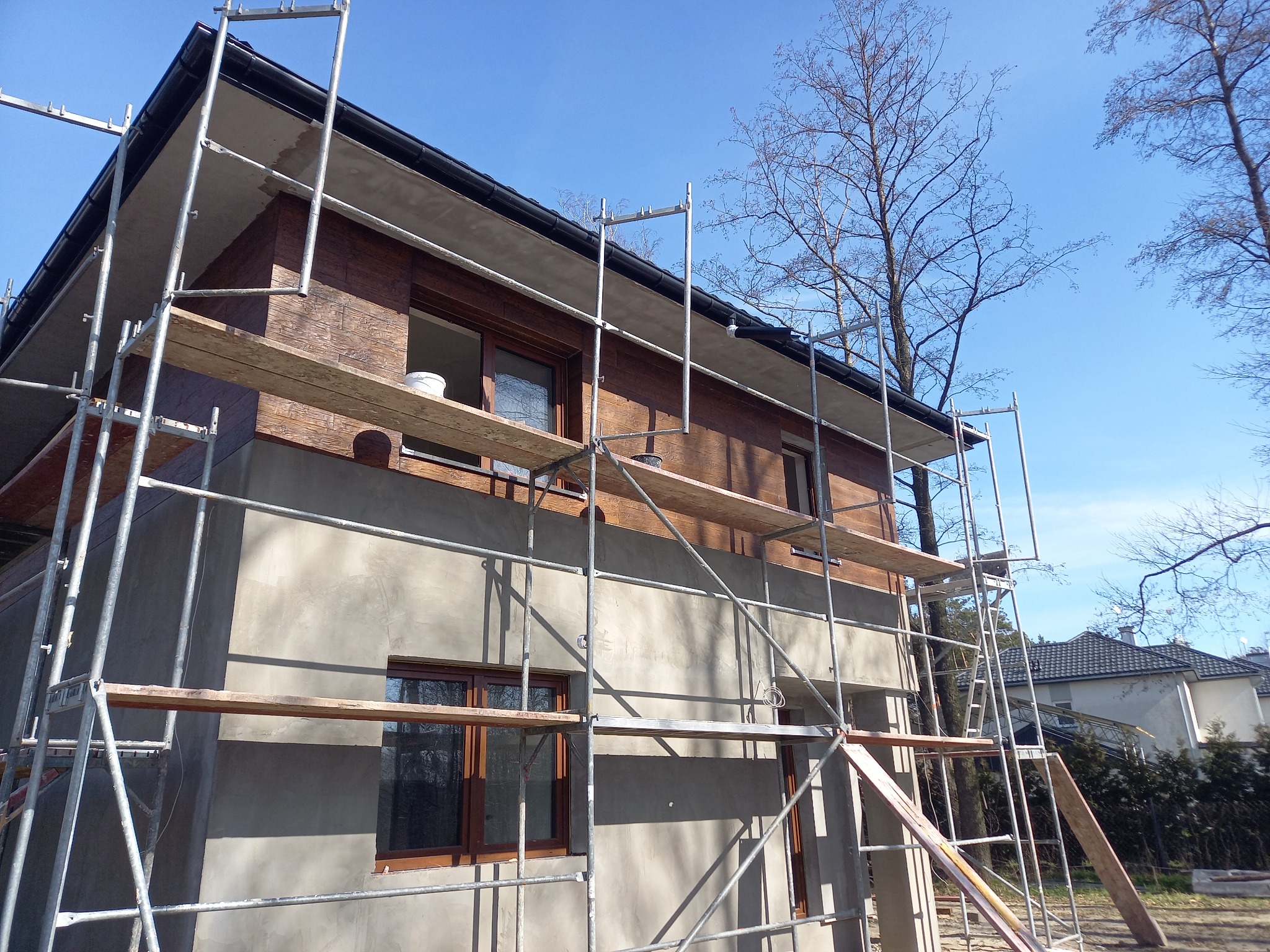 Dom jednorodzinny w trakcie prac elewacyjnych; dolna część budynku pokryta szarym tynkiem, górna wykończona imitacją drewnianej okładziny; widoczne rusztowania i niebieskie niebo.