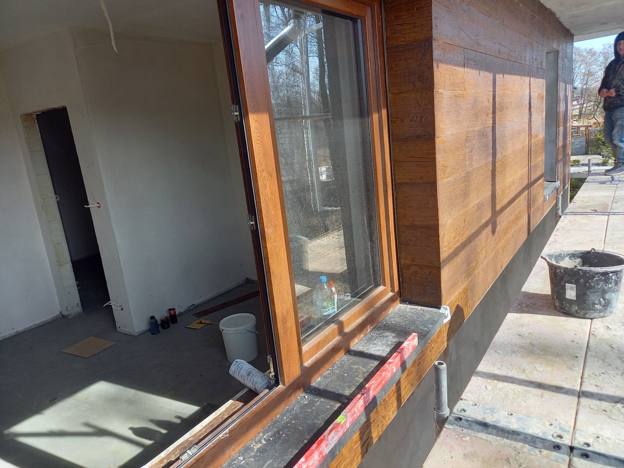 Elewacja budynku z drewnopodobnym wykończeniem, widoczne okno z otwartą ramą, poziomica na parapecie, wiadro i rusztowanie na zewnątrz, surowe wnętrze w tle.