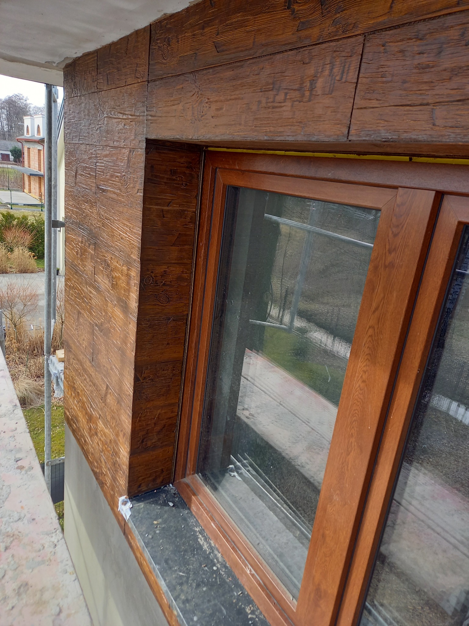 Elewacja budynku z oknem, wykończona panelami imitującymi drewno, widok z balkonu, w tle budynek mieszkalny i drzewa.