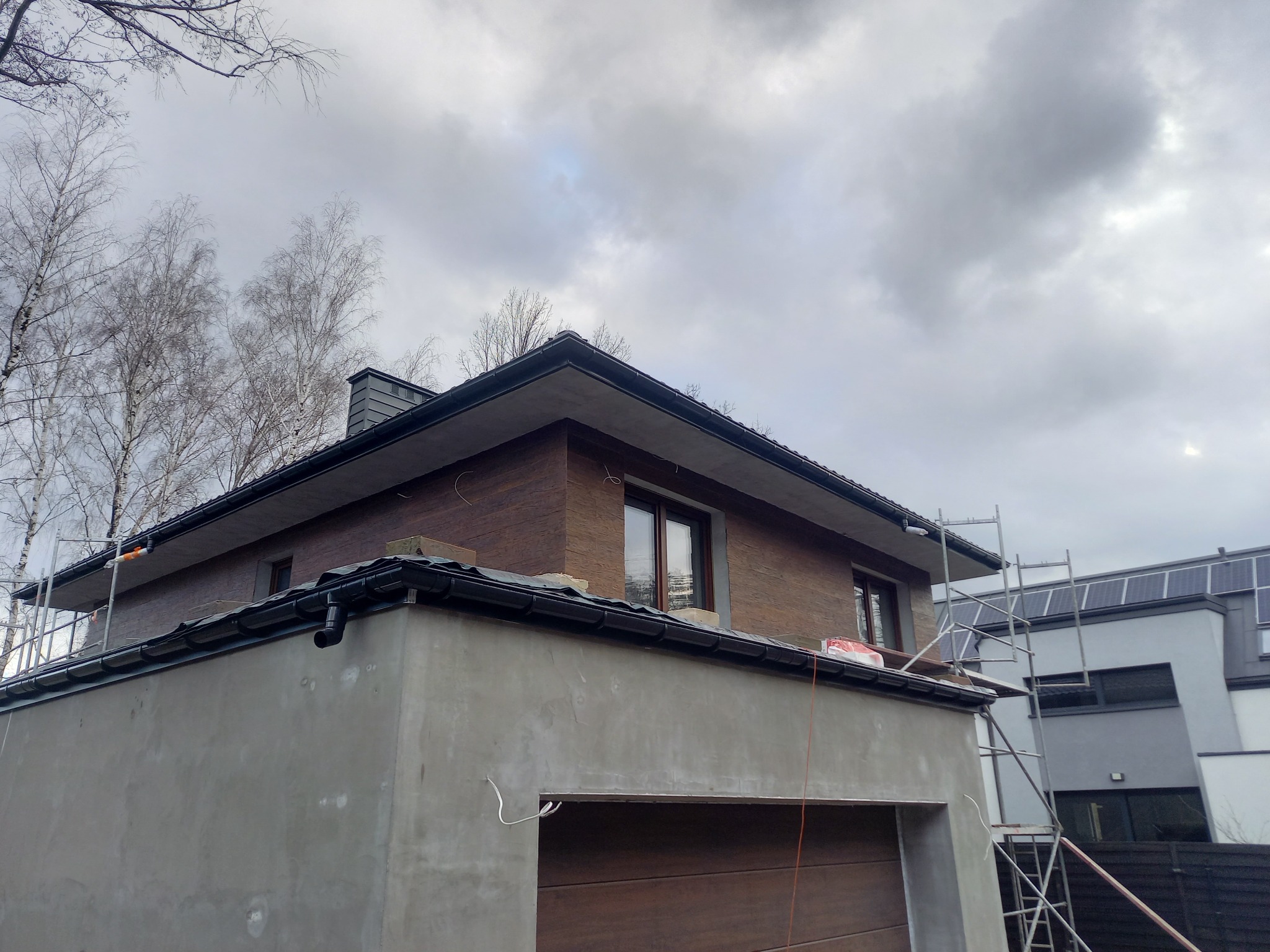 Surowa elewacja garażu połączona z drewnianą elewacją domu, widoczne rusztowania i orynnowanie, pochmurne niebo w tle.