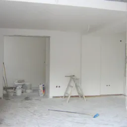 Wnętrze w trakcie remontu, widoczne świeżo pomalowane ściany na biało, drabina, narzędzia malarskie i worki z zaprawą, zarys toalety w przejściu do innego pomieszczenia, okno z luksferów.