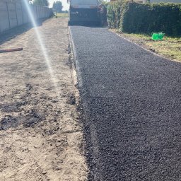 Wiśniewski €€€€€ - Układanie nowej nawierzchni asfaltowej na drodze dojazdowej, z widoczną maszyną rozściełającą i dwoma pracownikami w odblaskowych ubraniach, po jednej stronie gruntowa powierzchnia, po drugiej...