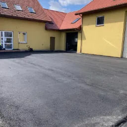 Nowa, gładka nawierzchnia asfaltowa przed budynkiem z żółtą elewacją i czerwoną dachówką, widoczna zagęszczarka w tle.