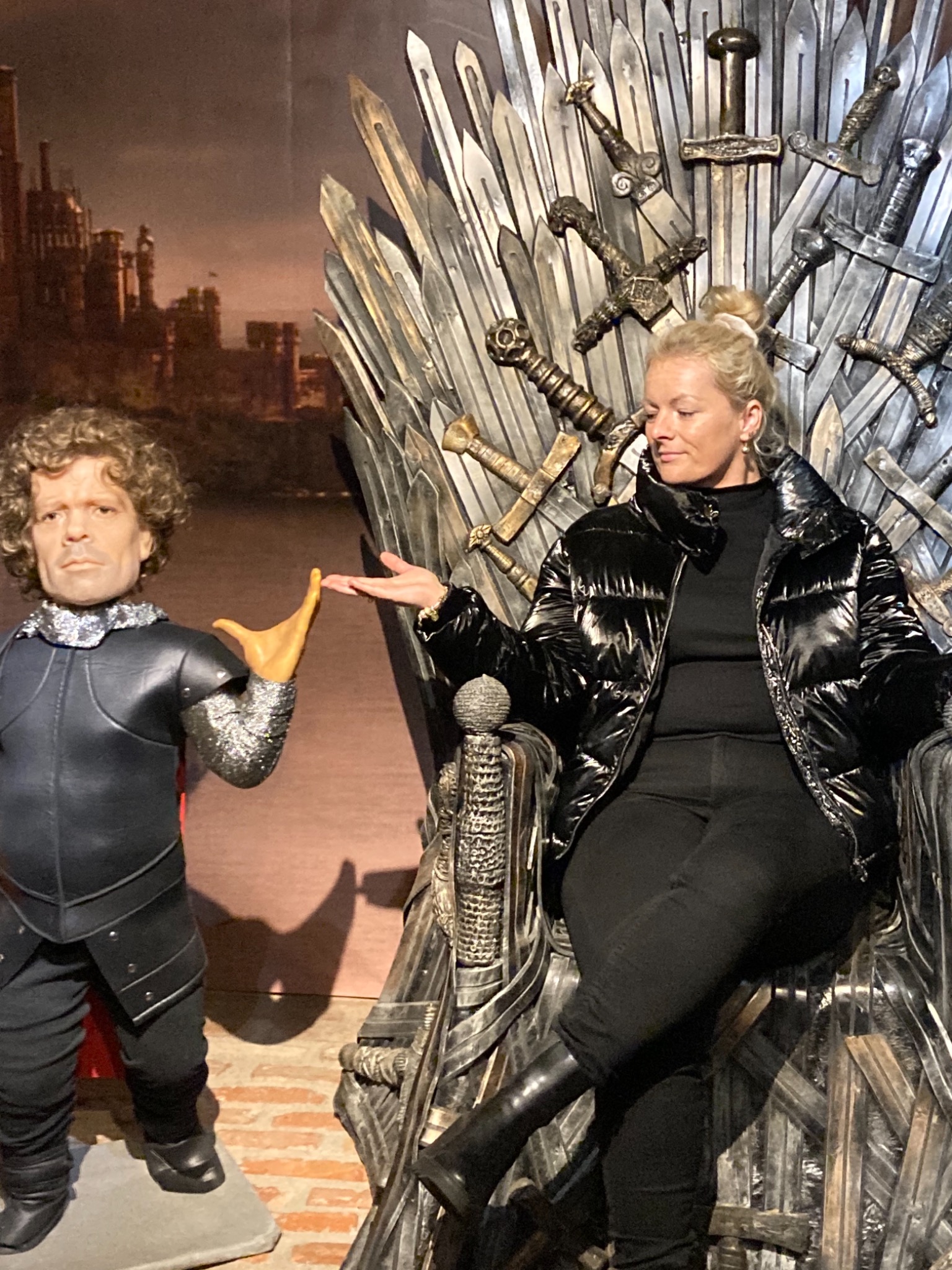 Kobieta w czarnej kurtce puchowej siedzi na tronie z mieczy, obok niej stoi figura Tyriona Lannistera z serialu Gra o Tron, w tle widoczna panorama miasta.