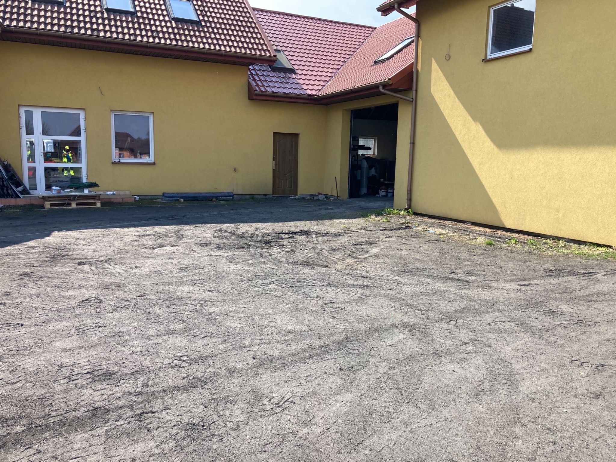 Przygotowany teren pod budowę z widocznymi śladami opon, żółty budynek z czerwonym dachem w tle.