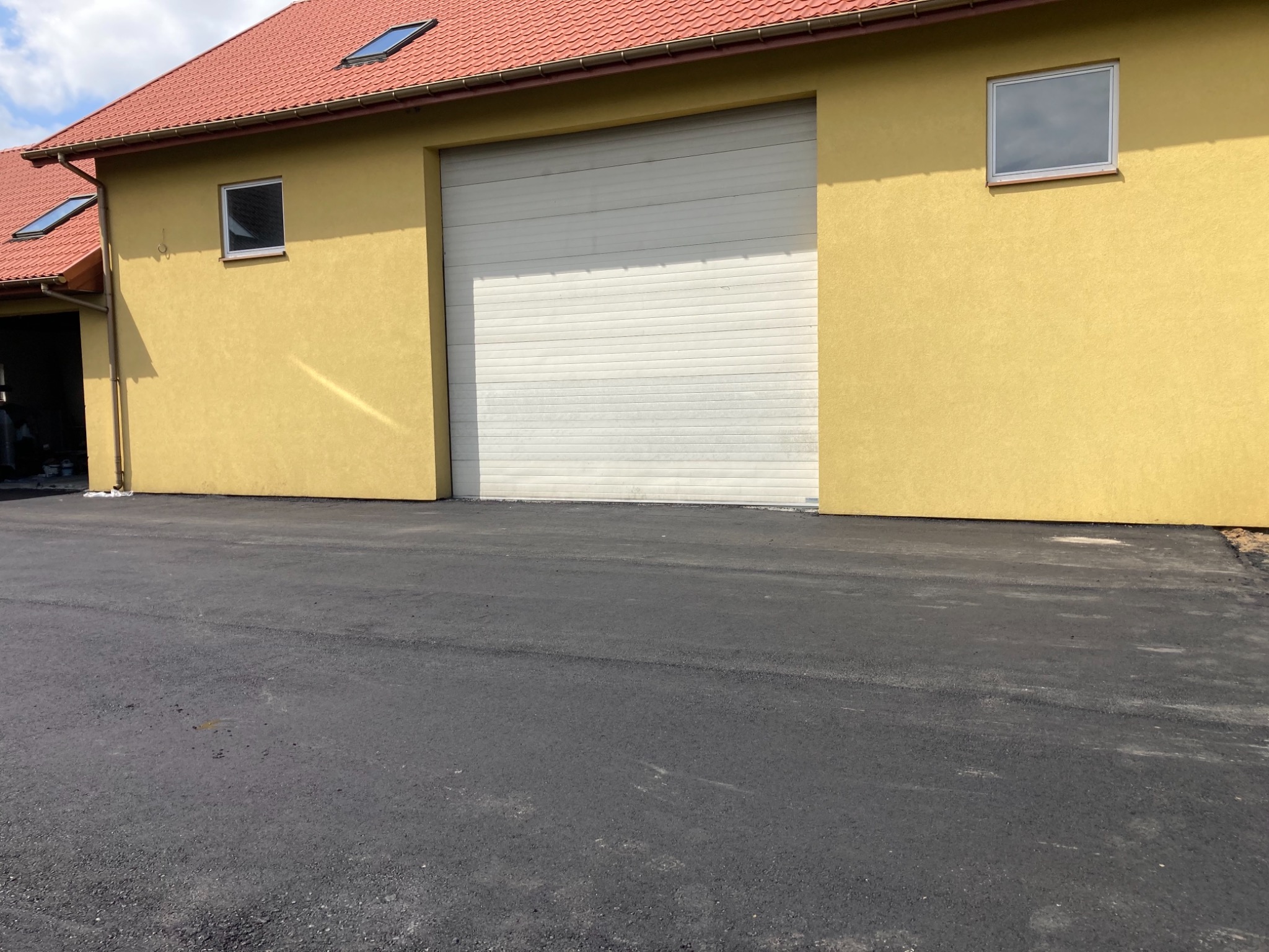 Żółty budynek gospodarczy z bramą garażową i nową, czarną nawierzchnią asfaltową przed nim.