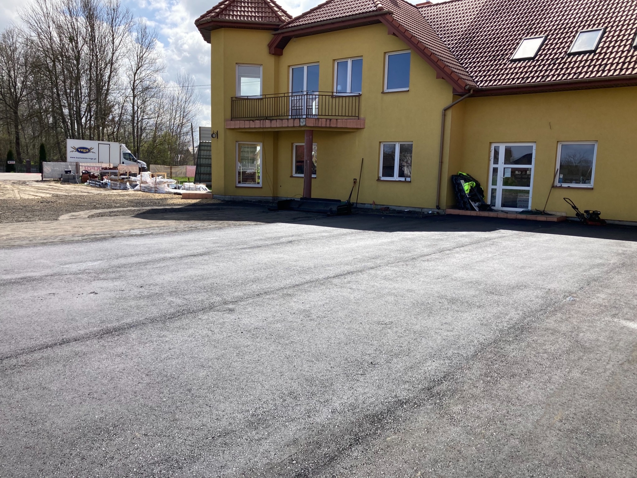 Nowa, świeżo położona nawierzchnia asfaltowa przed dwupiętrowym budynkiem z balkonem, widoczny sprzęt budowlany i dostawczy bus na posesji.