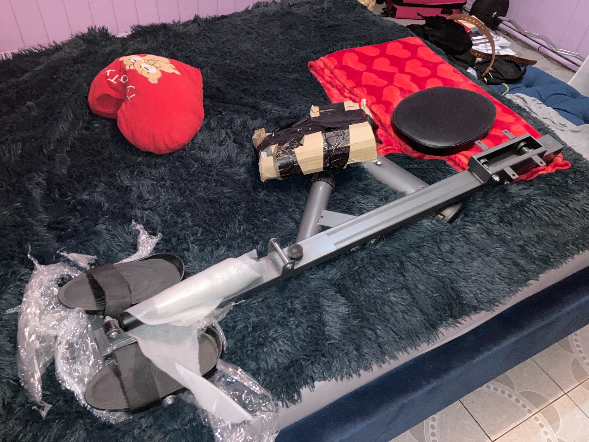 Rozpakowany, częściowo zmontowany sprzęt do ćwiczeń (ławka) leży na puszystym, ciemnym dywanie, obok poduszki w kształcie serca, zapakowanego elementu i pary klapek.