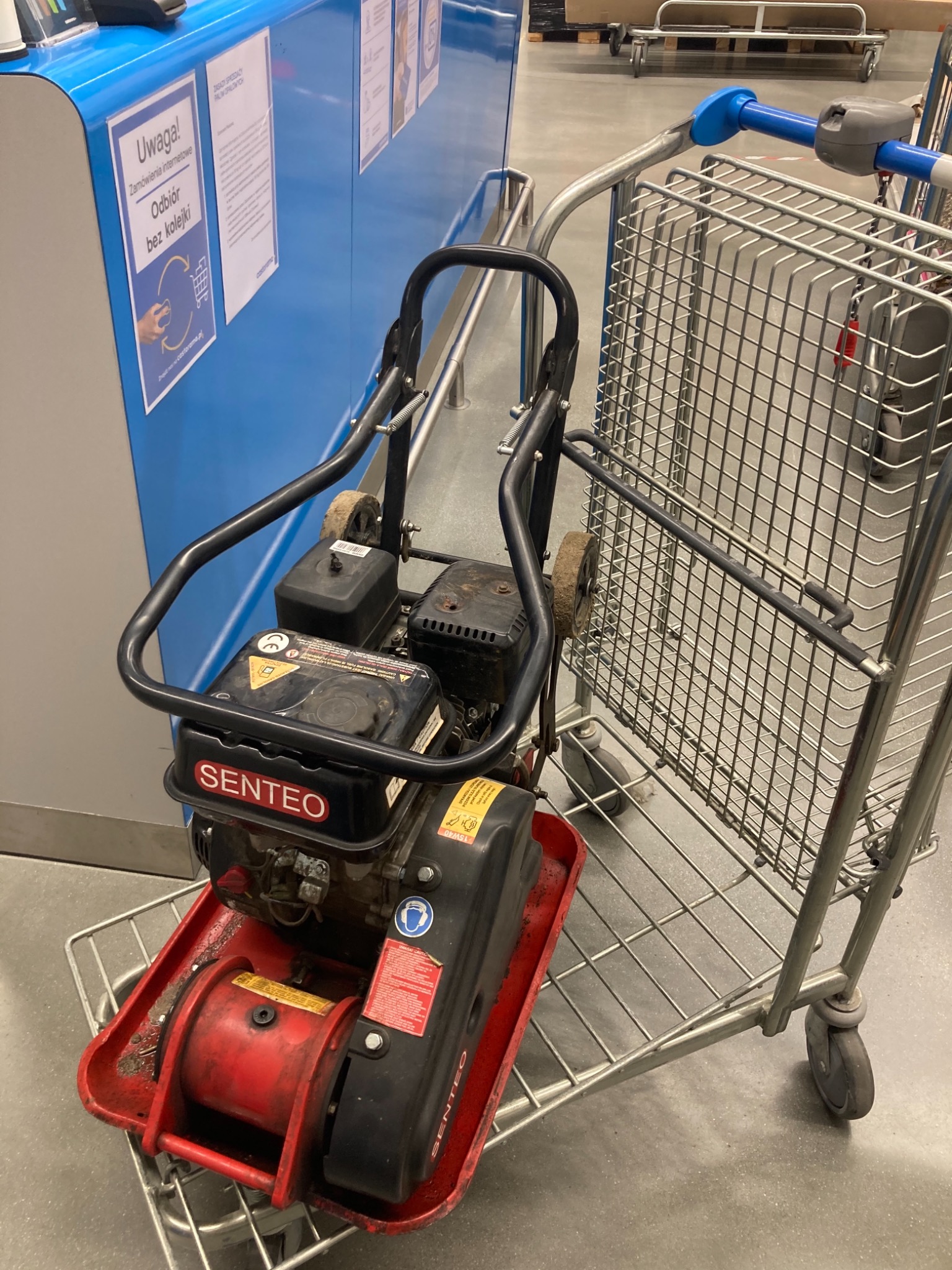 Zagęszczarka gruntu marki Senteo umieszczona w metalowym wózku sklepowym na tle kasy samoobsługowej.