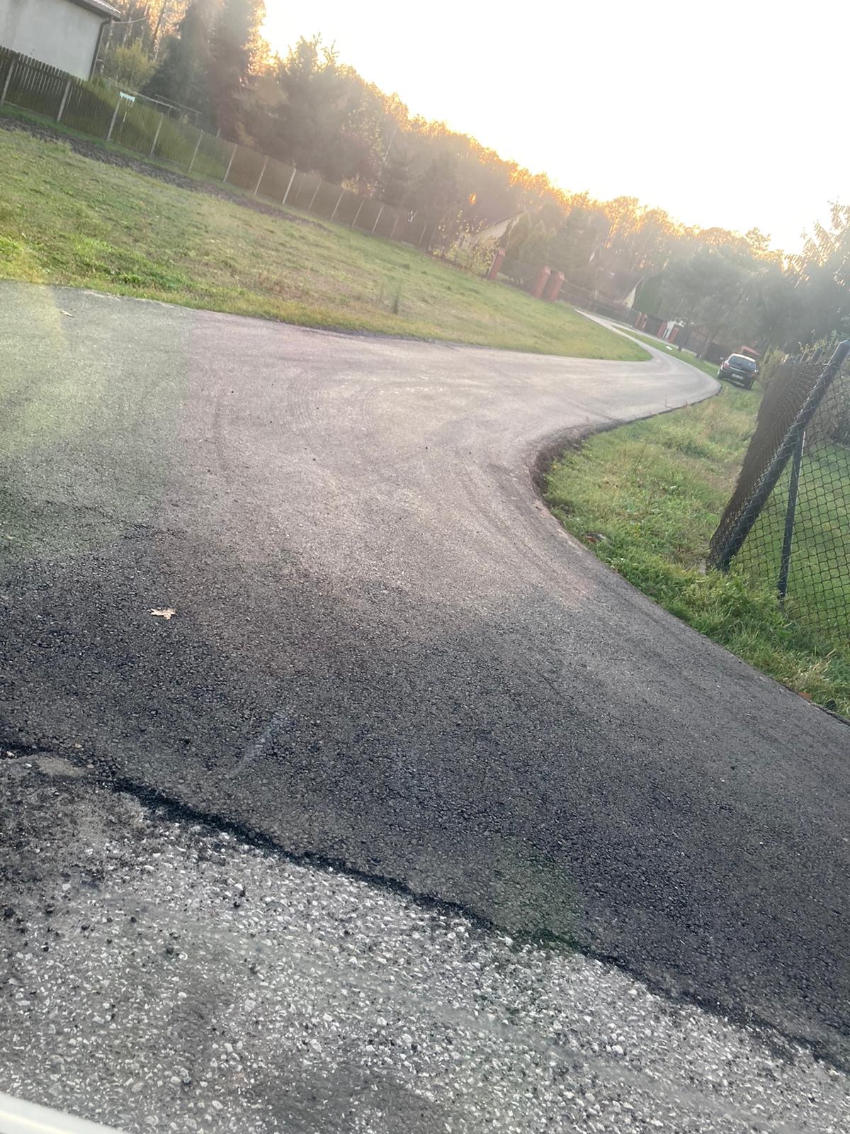 Nowo położona, zakręcająca droga asfaltowa prowadzi przez teren z trawą i drzewami, widoczny samochód i ogrodzenie z siatki.