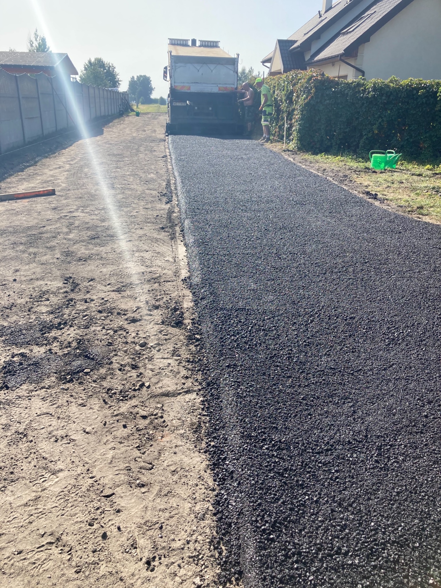 Układanie nowej nawierzchni asfaltowej na drodze dojazdowej, z widoczną maszyną rozściełającą i dwoma pracownikami w odblaskowych ubraniach, po jednej stronie gruntowa powierzchnia, po drugiej...