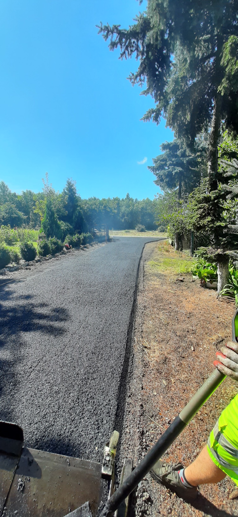 Układanie nowej nawierzchni asfaltowej na drodze dojazdowej, widok z perspektywy operatora maszyny, zieleń w tle.