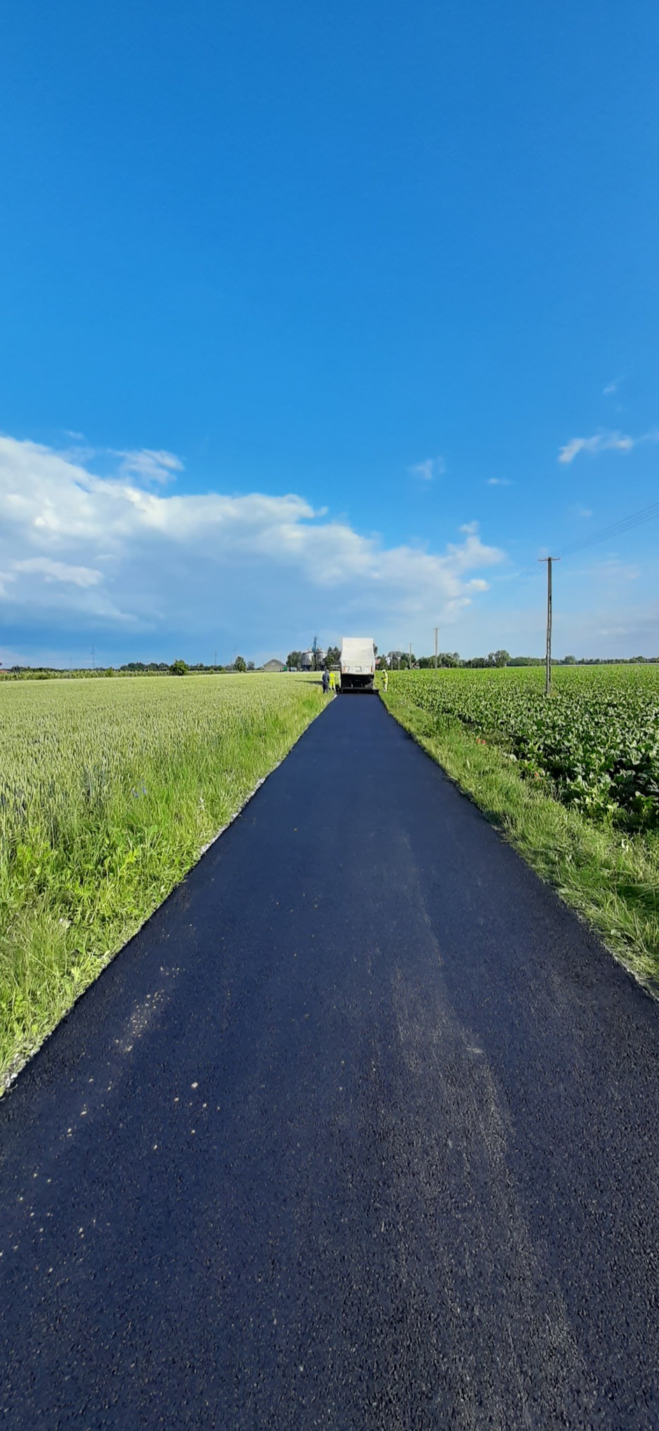 Świeżo ułożona, czarna nawierzchnia drogi biegnąca przez pola w słoneczny dzień, z widocznym sprzętem i pracownikami w oddali.