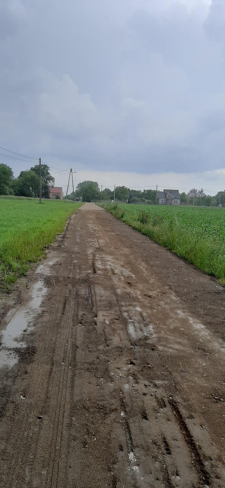 Ujęcie z perspektywy drogi gruntowej z koleinami po ciężkim sprzęcie, prowadzącej przez teren budowy w kierunku zabudowań wiejskich w pochmurny dzień.