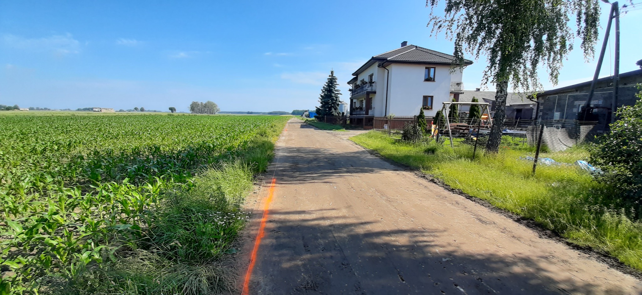 Przygotowany pas terenu pod budowę drogi na wsi, wytyczony pomarańczową farbą, obok pole kukurydzy i dom.