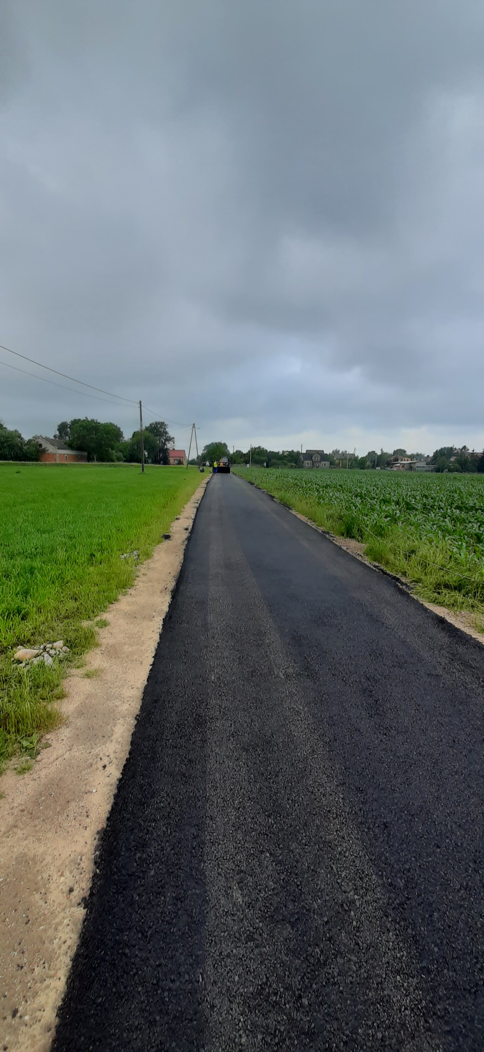 Świeżo położona czarna asfaltowa droga biegnąca przez wiejski krajobraz, z zielonymi polami po bokach i pochmurnym niebem w tle.