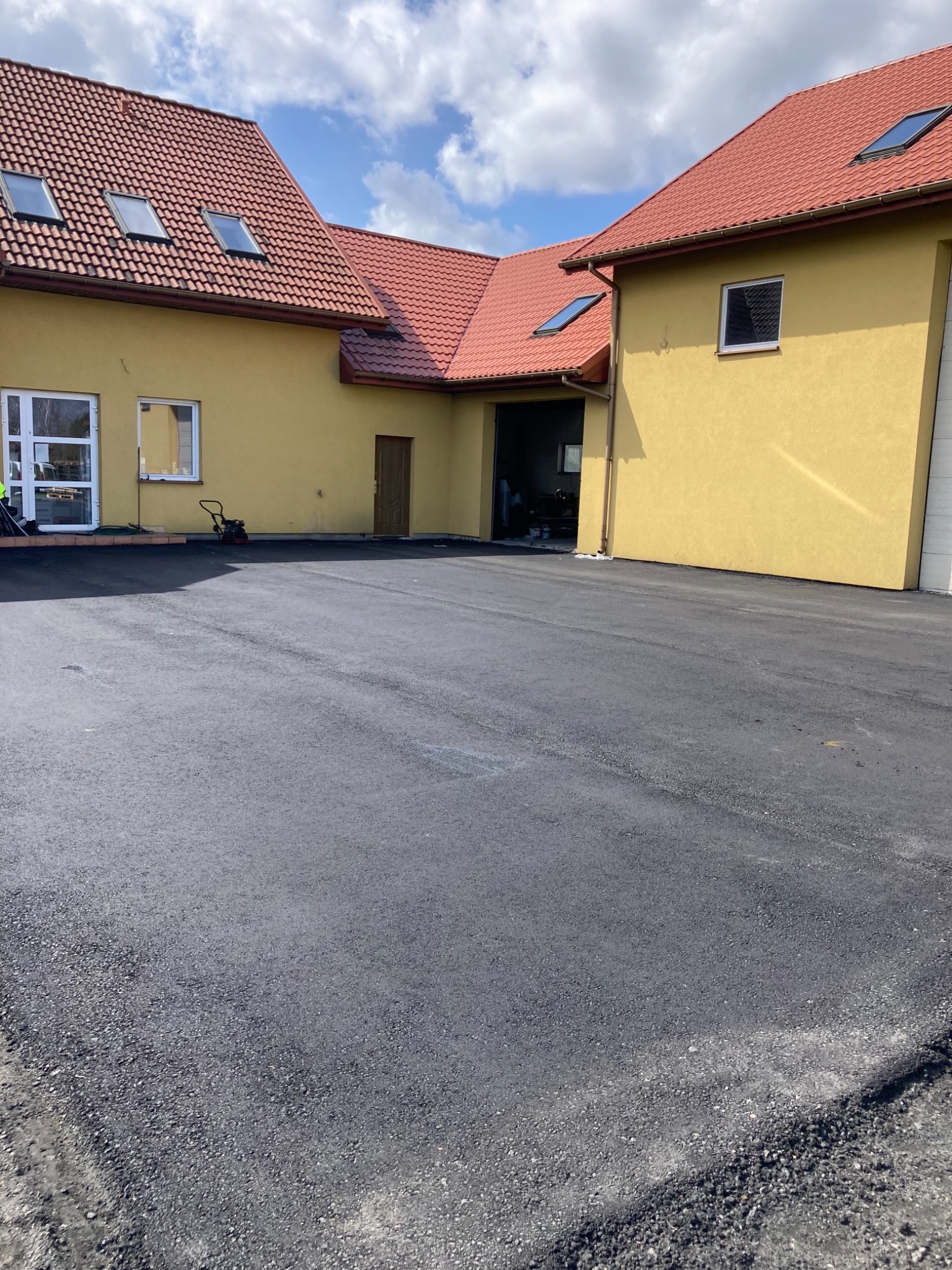 Nowa, gładka nawierzchnia asfaltowa przed budynkiem z żółtą elewacją i czerwoną dachówką, widoczna zagęszczarka w tle.