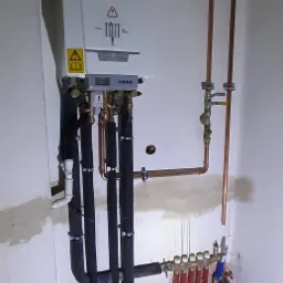 Biały piec grzewczy z podłączonymi rurami miedzianymi i czarnymi izolowanymi przewodami, widoczny rozdzielacz z kolorowymi rurami w nowym budynku w trakcie instalacji.