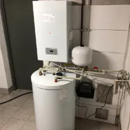 Biały, wiszący piec gazowy z podłączonym zasobnikiem ciepłej wody i instalacją, widoczne rury i zawory, na ścianie płytki ceramiczne.