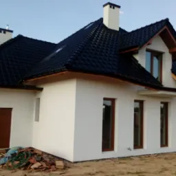 Nowo wybudowany dom jednorodzinny z ciemną dachówką i białymi ścianami, widoczne pozostałości materiałów budowlanych przed wejściem.