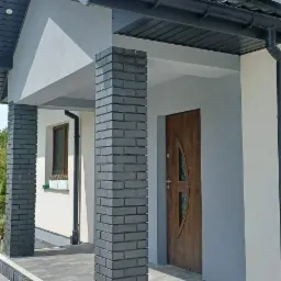 Nowoczesne wejście do domu z szarymi ceglanymi kolumnami, drewnianymi drzwiami z owalnym przeszkleniem i schodami wyłożonymi szarą kostką brukową, oświetlone naturalnym światłem słonecznym.