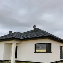 Nowoczesny dom jednorodzinny z białą elewacją, grafitową dachówką i czarnymi roletami zewnętrznymi, antena satelitarna na kominie, pochmurne niebo.