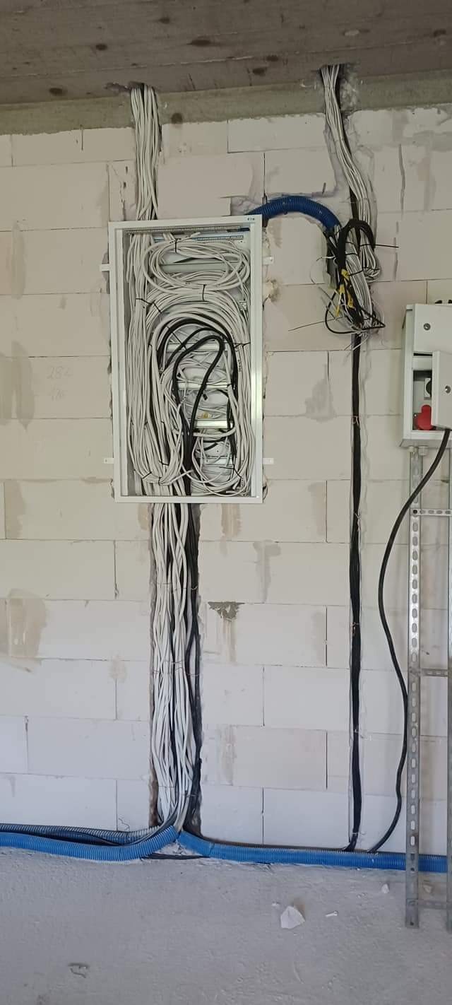 Rozdzielnica elektryczna z licznymi przewodami w białej obudowie, zamontowana na surowej ścianie z betonu komórkowego, widoczne rury karbowane w kolorze niebieskim i drabinka monterska.