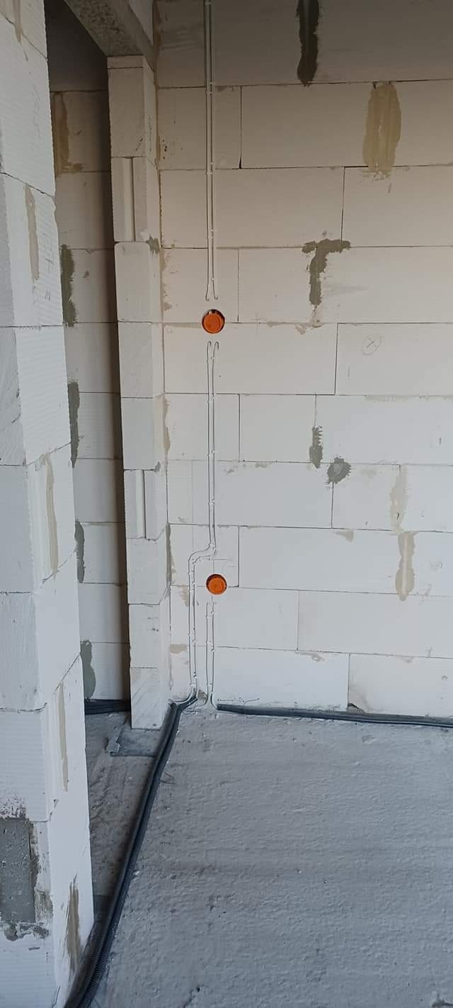 Surowe ściany z betonu komórkowego z widocznymi bruzdami pod instalację elektryczną i pomarańczowymi puszkami pod gniazdka, szare rury kablowe na podłodze.