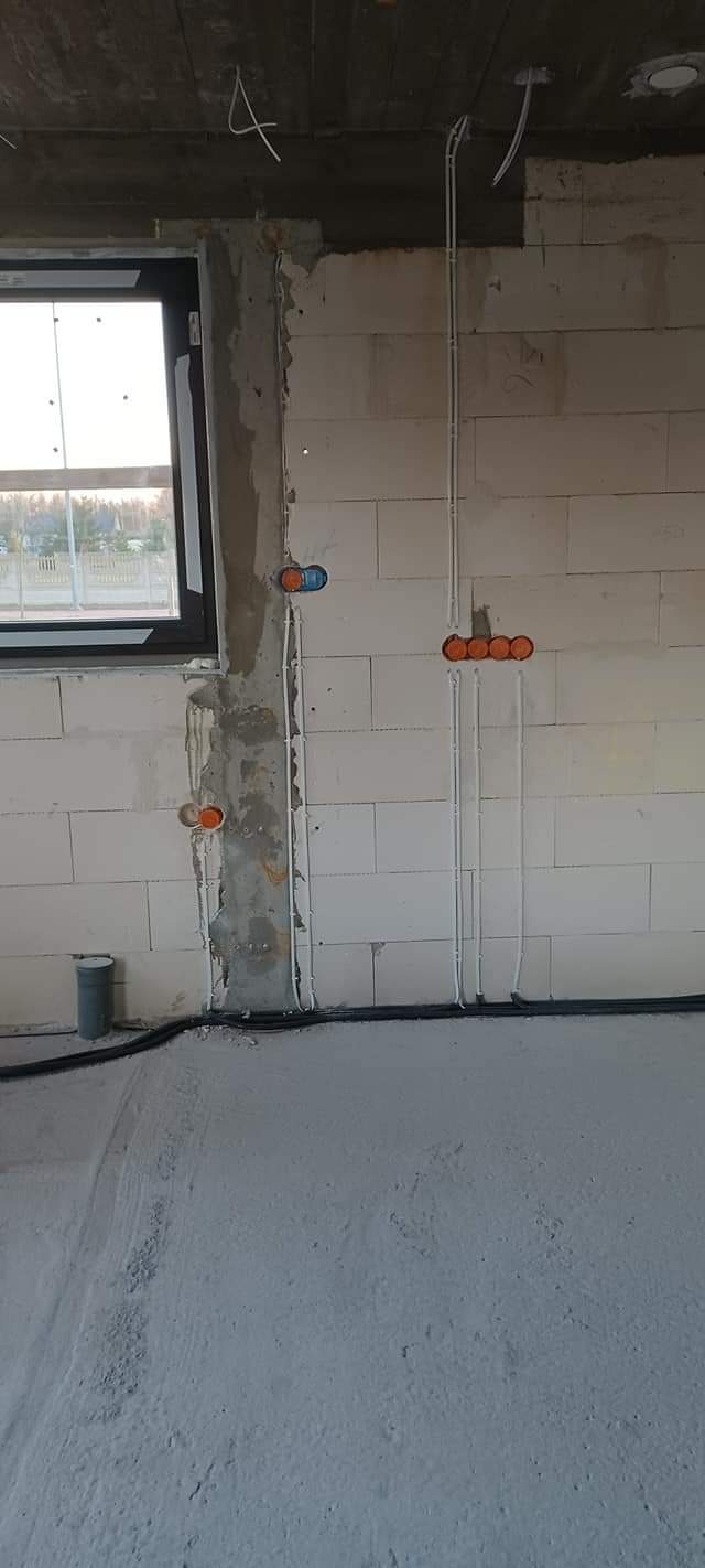 Surowy stan wnętrza w trakcie prac elektrycznych: widoczne kable, puszki instalacyjne w ścianie z betonu komórkowego, okno z widokiem na zewnątrz, rury przy podłodze, surowy betonowy sufit.
