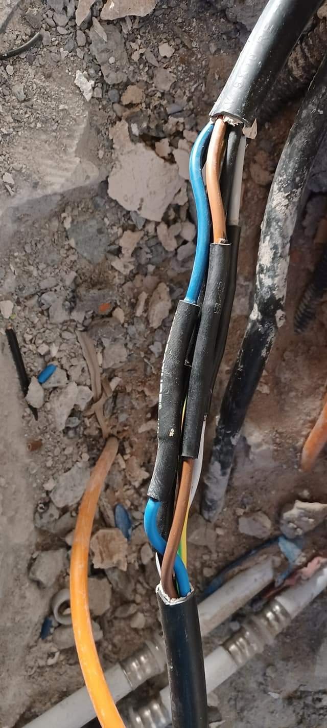 Połączenie przewodów elektrycznych z użyciem czarnej taśmy izolacyjnej, widoczne kolory izolacji: niebieski, brązowy, żółto-zielony, szary, w tle gruz i inne przewody.