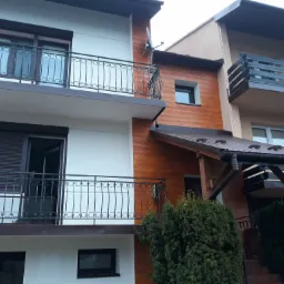 Dwupiętrowy budynek z białymi ścianami i drewnianą elewacją, balkony z ozdobnymi, metalowymi balustradami, widoczne okna z roletami zewnętrznymi.