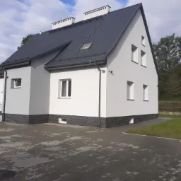 Nowoczesny, pomalowany na szaro-biało dom jednorodzinny w Humniskach z ciemną, metalową dachówką i ozdobnym ceglanym cokołem, widziany z zewnątrz na tle zieleni i błękitnego nieba.