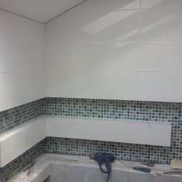 J.S.Project - Fragment łazienki w trakcie prac wykończeniowych, widoczne białe płytki na górze i mozaika z niebiesko-zielonych płytek na dole, z poziomym pasem białych płytek na środku, oraz narzędzia...