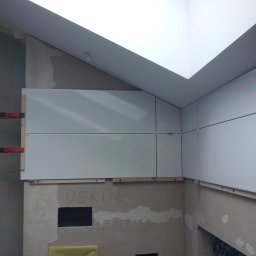 J.S.Project - Montaż białych płytek w narożniku pomieszczenia, widoczne drewniane listwy pomocnicze i zaciski, surowa ściana z napisem 'DEKOR'.
