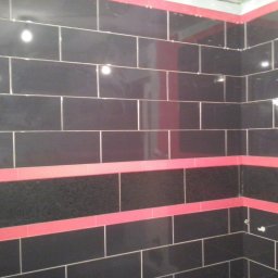J.S.Project - Ściana łazienki wyłożona błyszczącymi, czarnymi płytkami z poziomymi paskami czerwonych płytek dekoracyjnych i wzorzystych płytek, widoczne krzyżyki dystansowe.