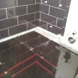 J.S.Project - Układanie czarnych, błyszczących płytek podłogowych z czerwonymi akcentami w łazience, widoczne krzyżyki dystansowe i fragment ściany z czarnymi płytkami ułożonymi w cegiełkę z czerwoną listwą...