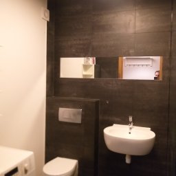 J.S.Project - Nowoczesna łazienka z ciemnymi płytkami, podwieszaną umywalką i toaletą, pralką z boku, oraz lustrem wnękowym odbijającym fragment innego pomieszczenia.