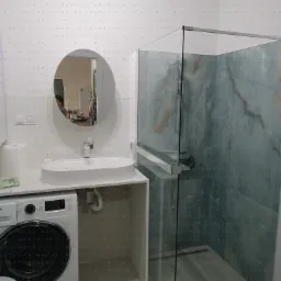 Nowoczesna łazienka z kabiną prysznicową typu walk-in, owalną umywalką nablatową i pralką w zabudowie. Minimalistyczny design, jasne barwy i marmurowe wykończenie ścian.