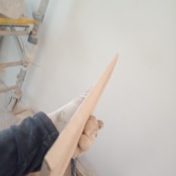 J.S.Project - Ręka w rękawicy trzyma listwę przypodłogową na tle świeżo pomalowanej ściany, widoczne rusztowanie i materiały budowlane.