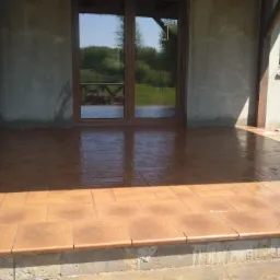 Brązowe, kwadratowe płytki ceramiczne na tarasie, świeżo po położeniu, odbijające światło, z widocznym fragmentem ściany budynku i narzędziami glazurniczymi w rogu.