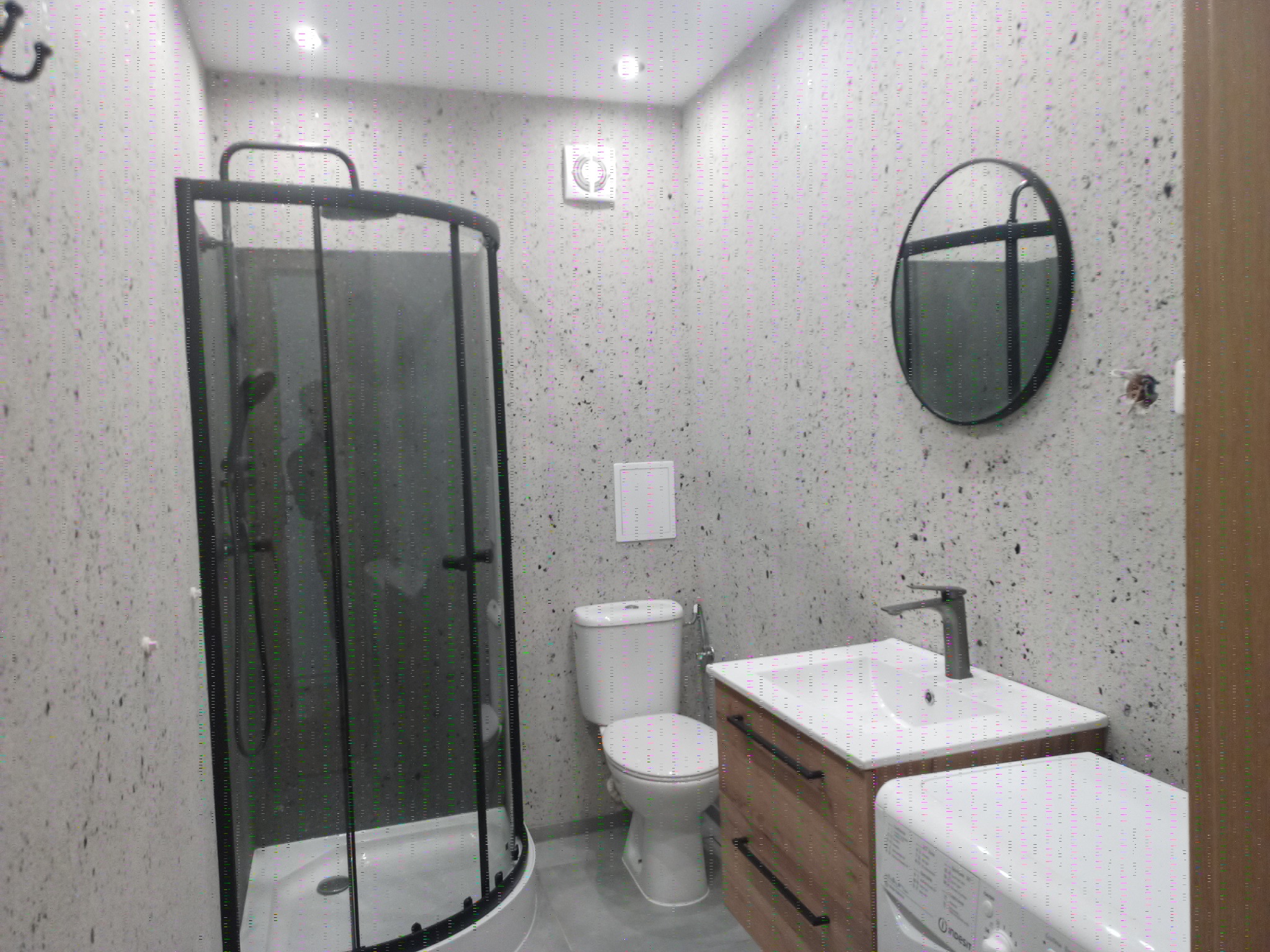 Nowoczesna łazienka z kabiną prysznicową, okrągłym lustrem i pralką, wykończona w szaro-białej kolorystyce z akcentami drewna. Minimalistyczny design.