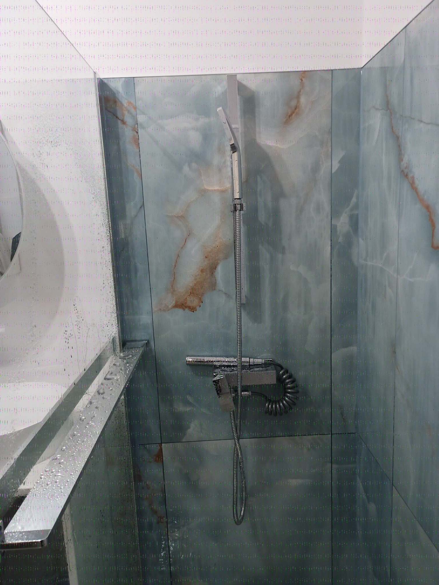 Nowoczesna kabina prysznicowa wykończona płytkami imitującymi marmur, z chromowaną armaturą i widocznymi kroplami wody na szklanych elementach.