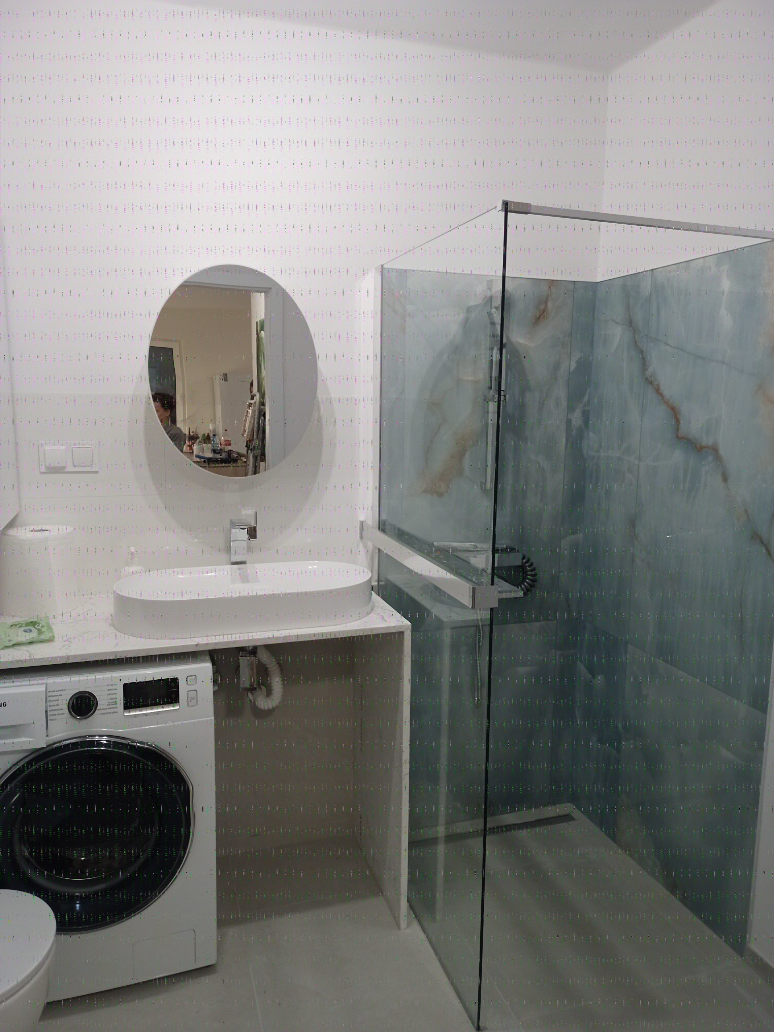 Nowoczesna łazienka z kabiną prysznicową typu walk-in, owalną umywalką nablatową i pralką w zabudowie. Minimalistyczny design, jasne barwy i marmurowe wykończenie ścian.