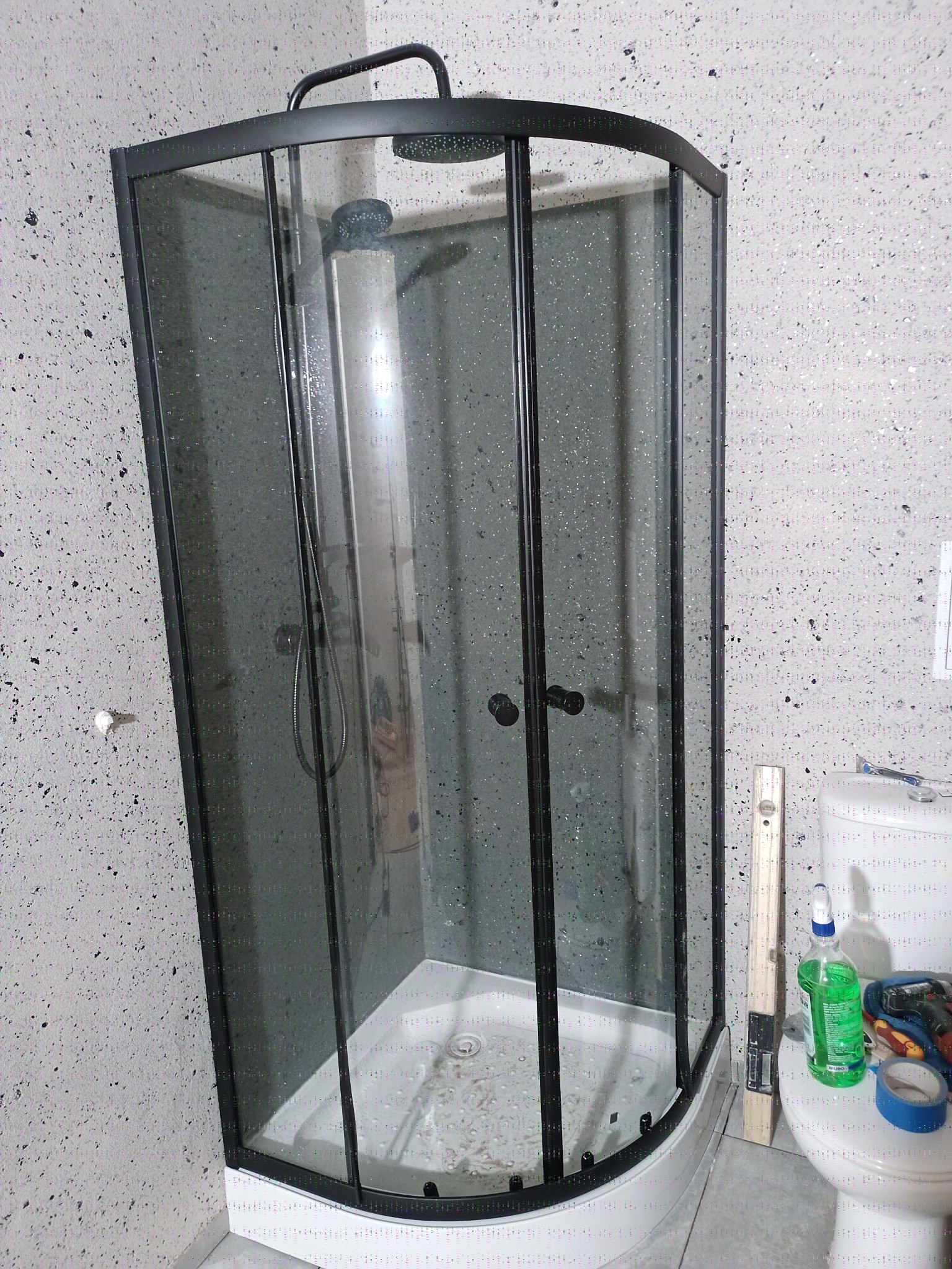 Nowoczesna kabina prysznicowa z czarnymi profilami na tle ściany w kropki. Widoczny poziomica i narzędzia, sugerujące świeży montaż w wykończonej łazience.