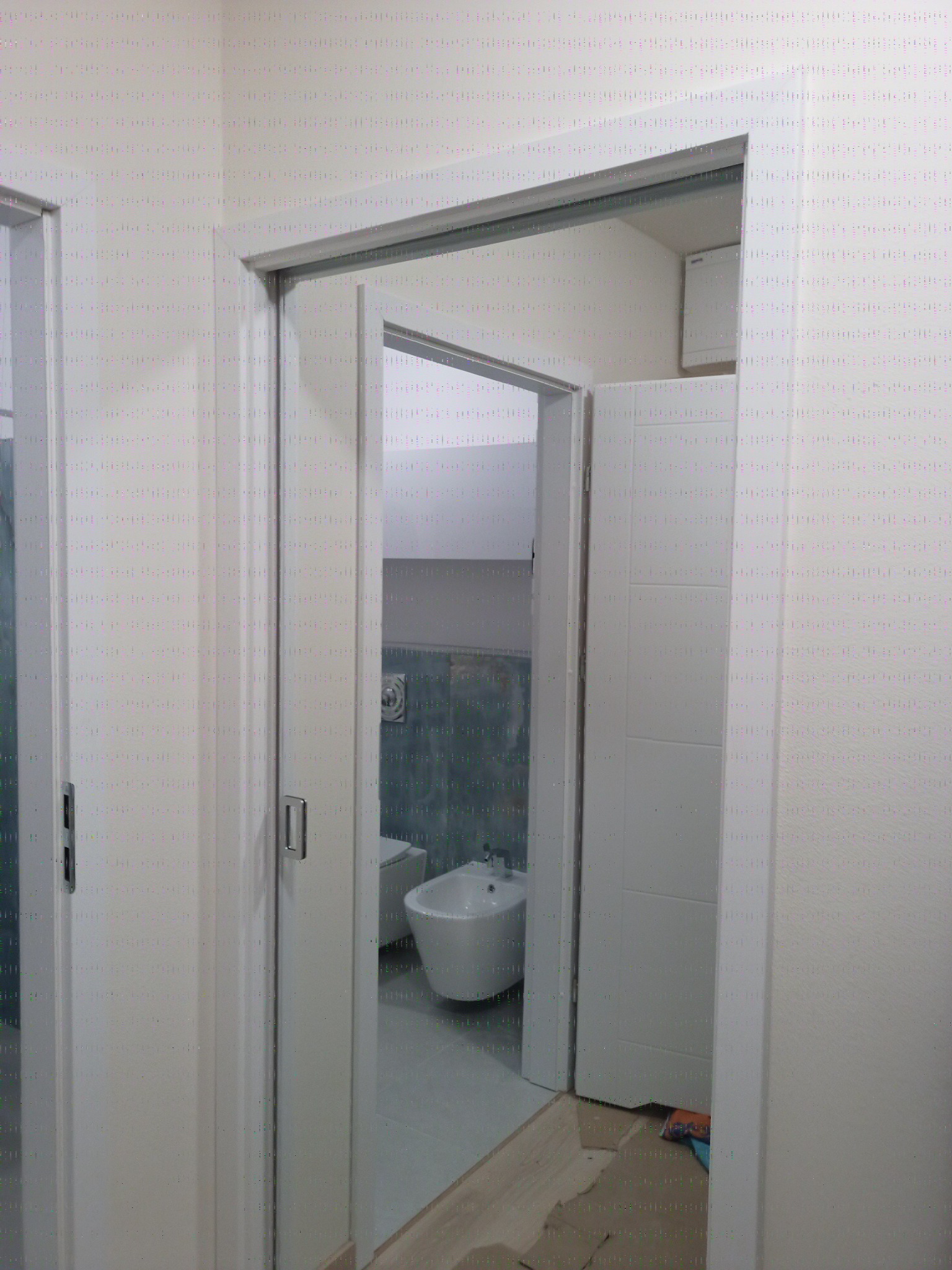 Nowoczesna łazienka po remoncie: białe drzwi przesuwne, bidet i toaleta z szarą mozaiką w tle. Widoczne zabezpieczenia podłogi podczas prac.