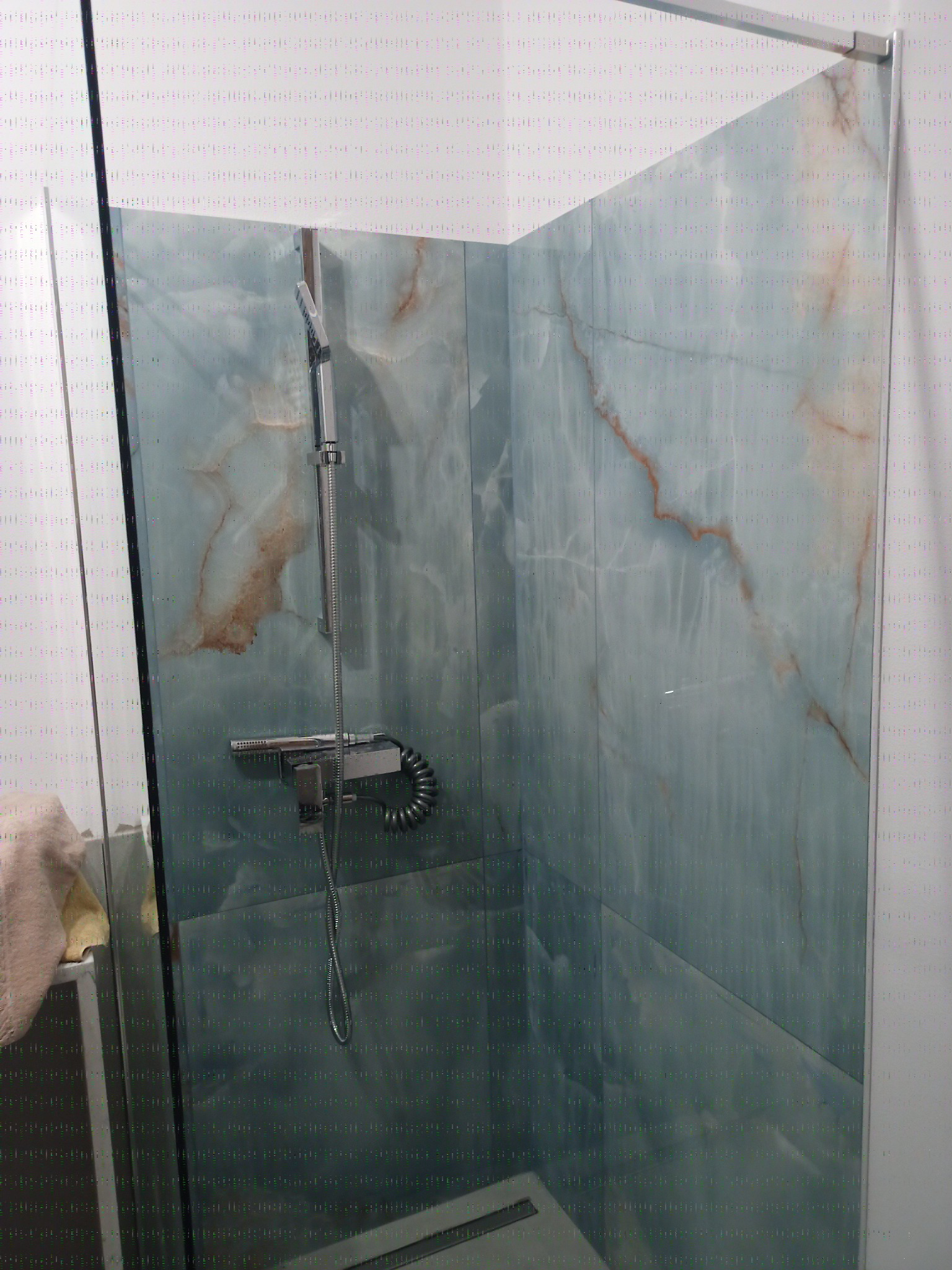 Szklana kabina prysznicowa z błękitnymi, marmurkowymi płytkami i chromowaną armaturą. Widoczny odpływ liniowy i fragment ręcznika.