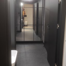 Grzegorz Smoleński - Nowoczesna toaleta publiczna z kabinami o czarnych drzwiach, szarą podłogą z płytek i okrągłymi lampami sufitowymi, widok z perspektywy umywalki z odbiciem osoby w lustrze.