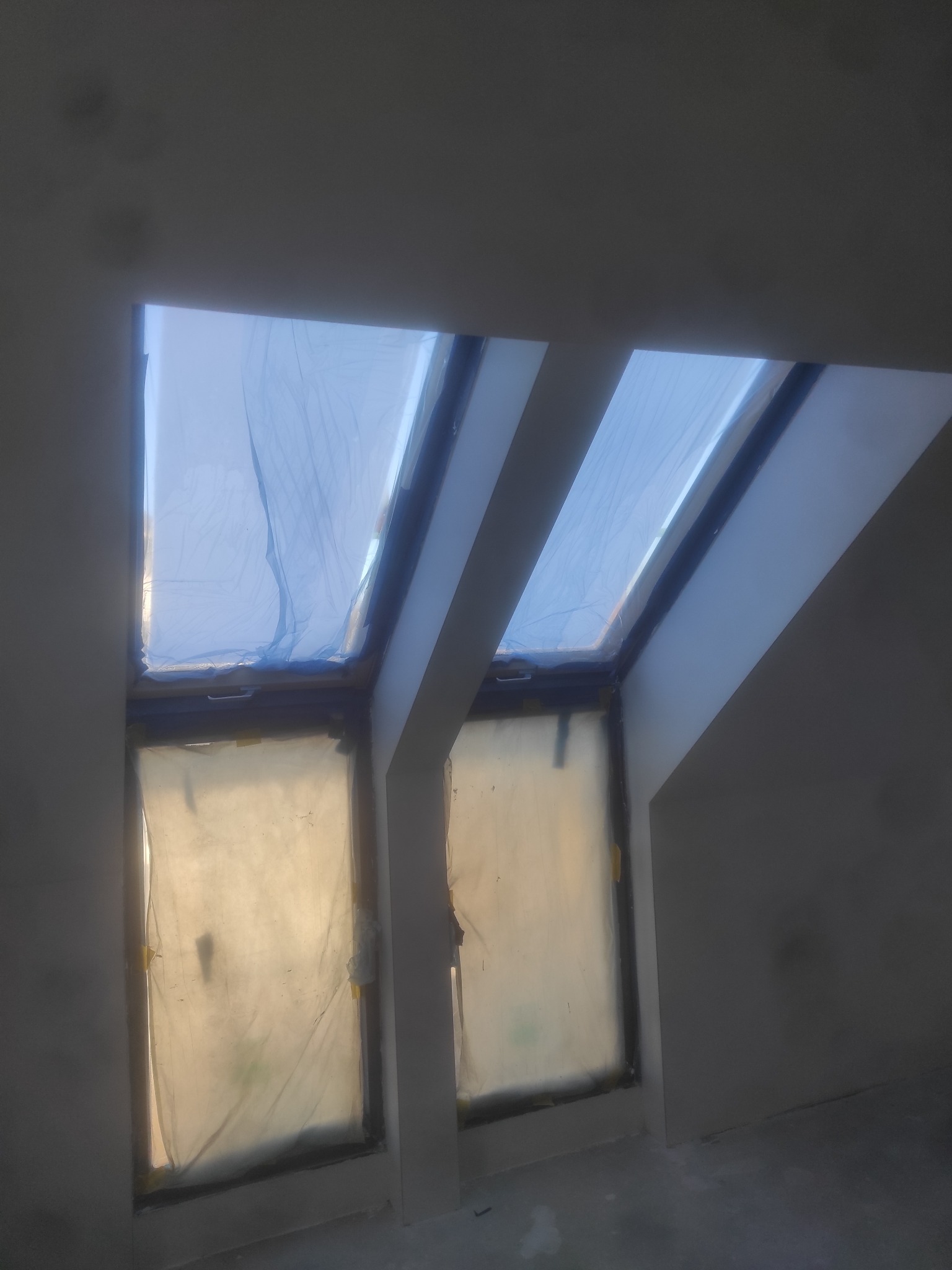 Dwa okna dachowe zabezpieczone folią malarską podczas prac wykończeniowych wnętrza na poddaszu.