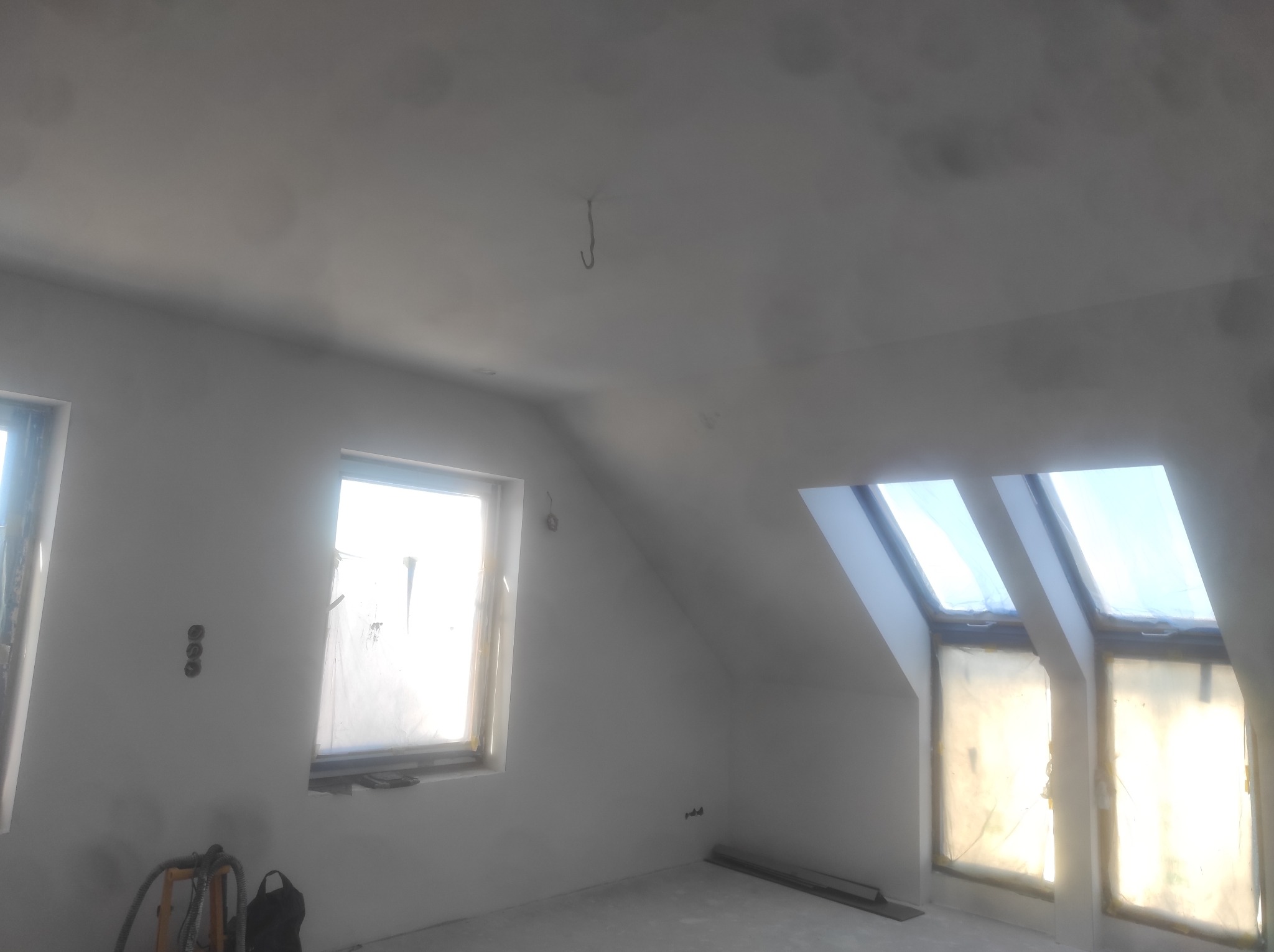 Poddasze w trakcie remontu z oknami dachowymi zabezpieczonymi folią, widoczne puszki elektryczne i wiszący hak na suficie.
