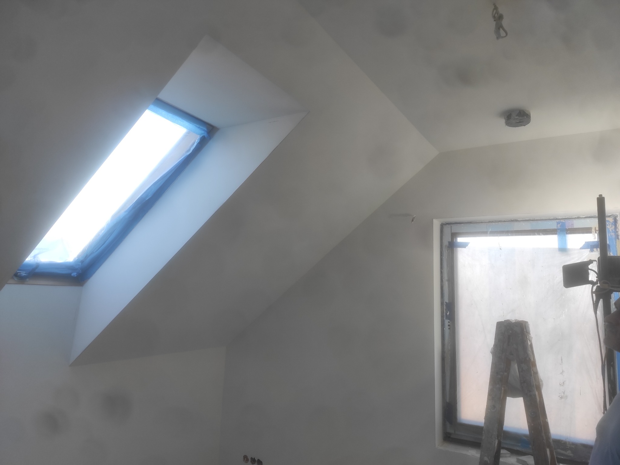 Poddasze po malowaniu: okno dachowe i zwykłe zabezpieczone folią malarską, widoczne puszki elektryczne w ścianie, drabina malarska przy oknie.
